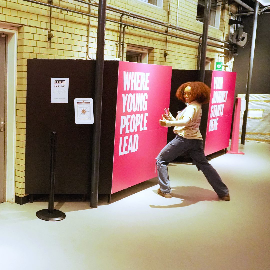 A female participant  stood in front of a pink poster with white text saying 'Where Young People Lead'.